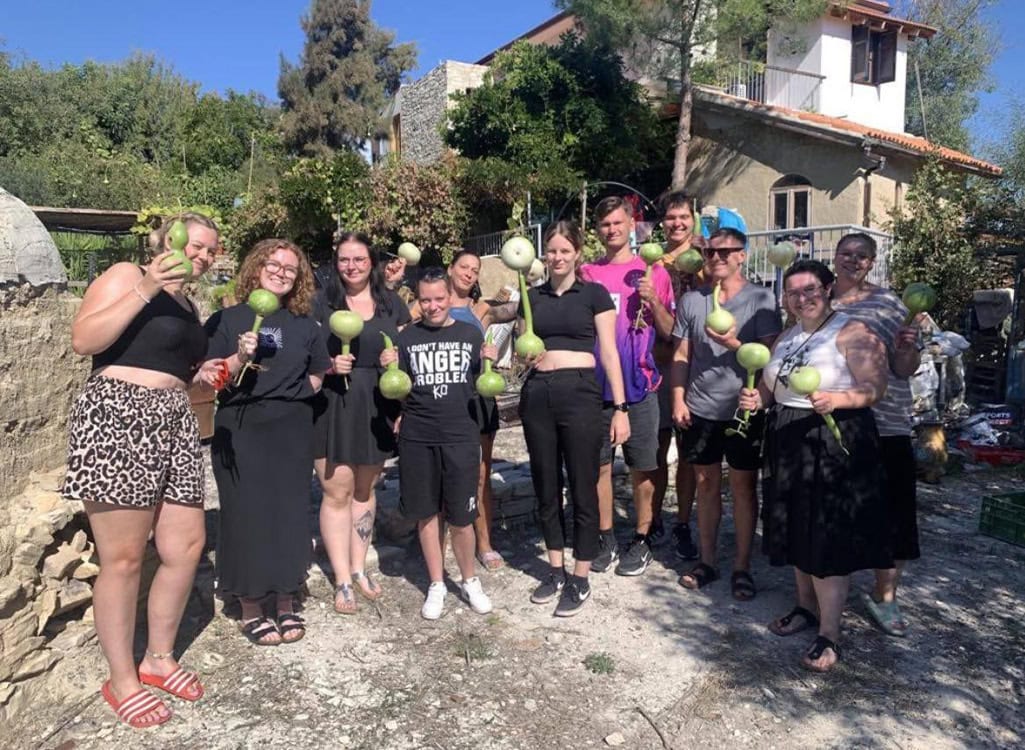 Gruppe von Menschen lächelnd, halten grüne Kürbisse, sonnig, vor einem alten Haus stehend.